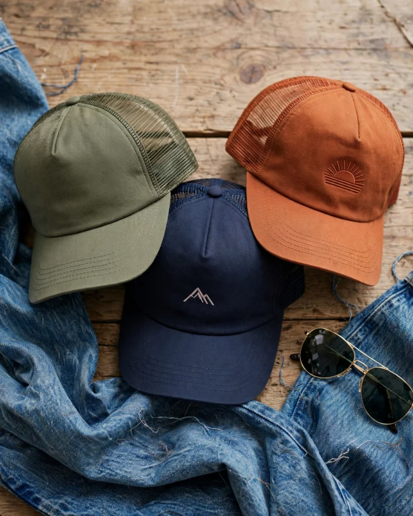 Three trucker caps arranged on a weathered wooden table. A heather grey cap with a circular embroidered patch on the front, a deep navy cap with a breathable mesh back, and a clean white cap with a blue mesh back panel. Each shows the characteristic curved brim and structured six-panel front. To the side: a folded piece of raw indigo denim, a pair of matte black wayfarer-style sunglasses, and a ceramic mug. The lighting is soft and overhead, casting gentle shadows that emphasize the texture of the cotton twill fabric.