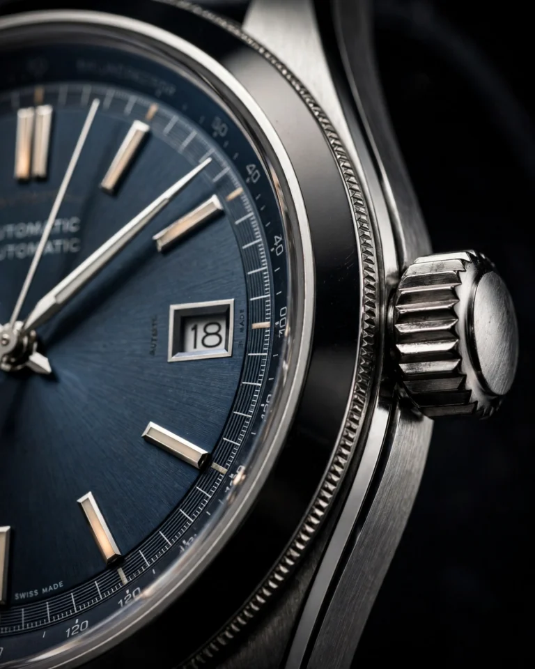 An extreme macro photograph of a minimalist wristwatch, focusing on the dial face and the crown. The white dial with slim hour markers and thin hands is visible in sharp detail, along with the polished stainless steel case and the textured crown at the three o'clock position. The milanese mesh band extends out of frame on both sides. Dramatic side lighting creates highlights on the polished metal and subtle shadows in the dial texture. The background is pure black, isolating the watch and emphasizing its precision-instrument quality. The overall mood is technical and reverent.